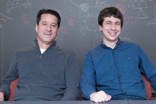Michael Kearns and Aaron Roth pose in front of an equation-filled chalkboard.