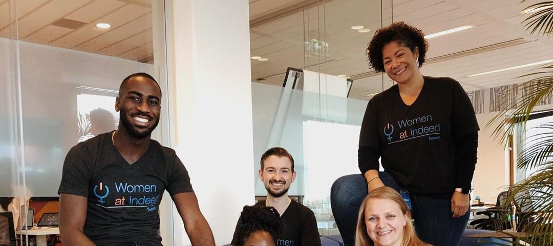 Image of five Indeed inclusion group members smiling and wearing shirts labeled “Women at Indeed” in a relaxed office setting