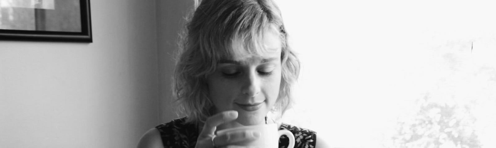 A black and white photo of a woman drinking a cup of coffee by a window in her home.