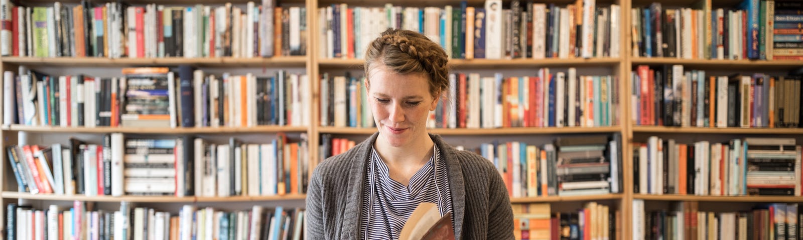 A young female customer  at bookstore standing against a bookshelf picks up a book and reads through it.