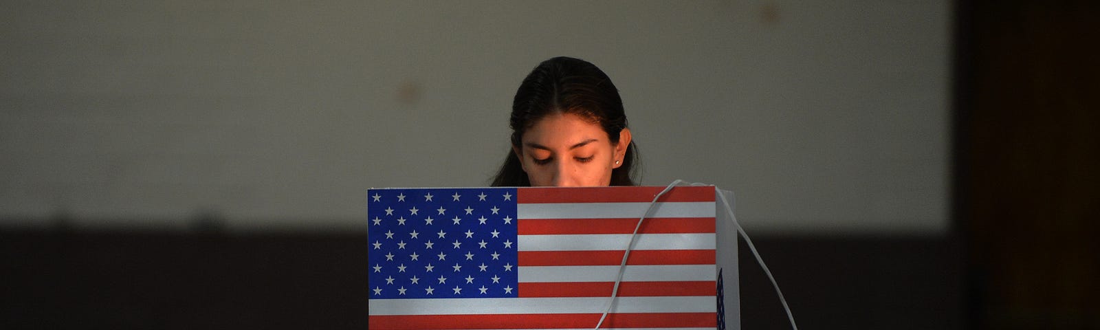 Latina woman voting at a poll in California. The booth has an American flag on it.