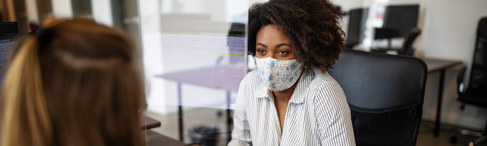 A Black businesswoman wearing a face mask negotiates with a client. There is a plastic divider for social distancing.