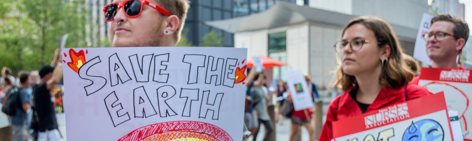 A group of protesters on a city sidewalk carrying signs that say “save the earth” and “hot earth summer”