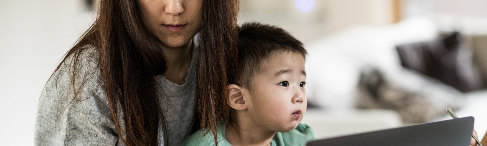 A photo of a mom working on her laptop while her son sits in her lap, she looks busy and tired.