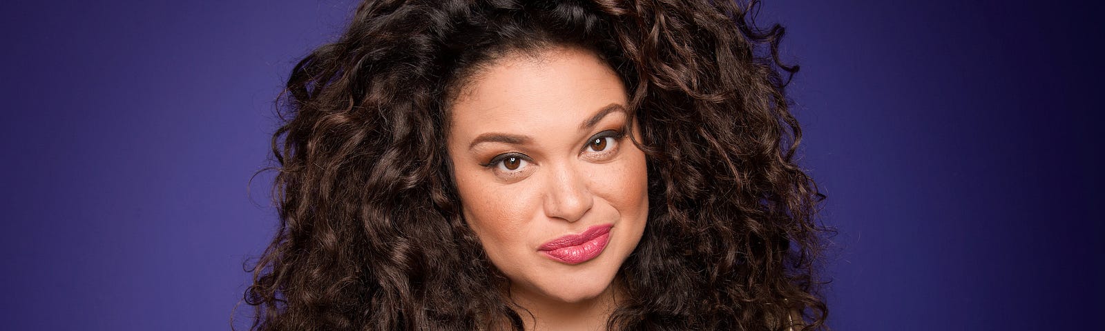 Portrait photo of Michelle Buteau in front of an indigo background.