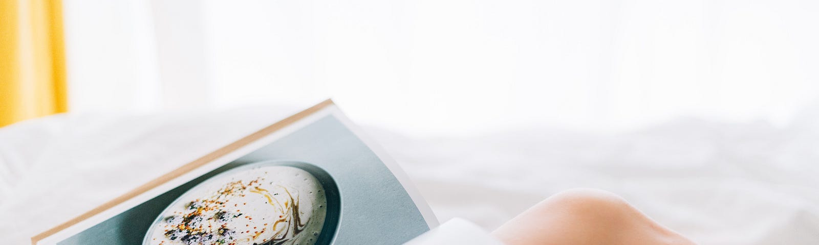 Closeup of a person, from the neck down, who’s reclining in bed, reading a cookbook with a large photo on one page.