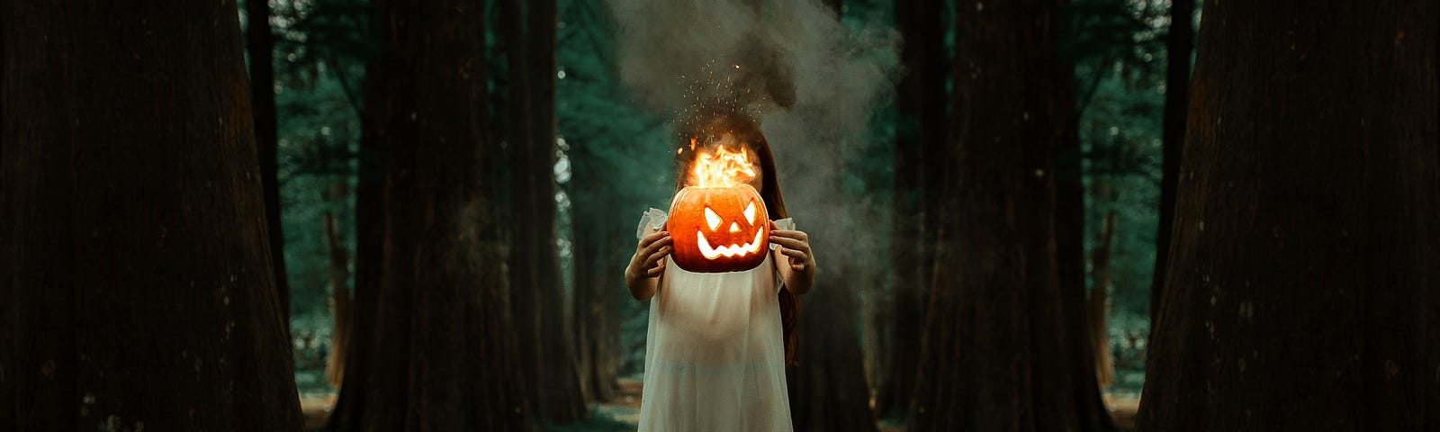 A girl wearing a white dress is standing in the middle of a forest. She’s holding a burning (carved) pumpkin in front of her face.