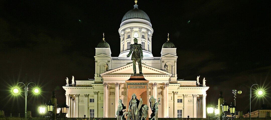 A night time scene of a grand, floodlit, white building with a statue of a man standing above other figures at the base of the monument in the foreground. The words, ‘Emperor Alexander II of Russia, Grand Duke of Finland’ are superimposed at the bottom of the image.