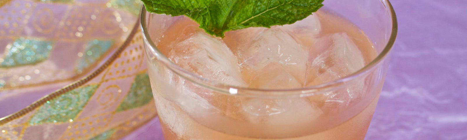 A cloudy, refreshing ginger-colored drink on ice, with a mint sprig and a lavender backdrop