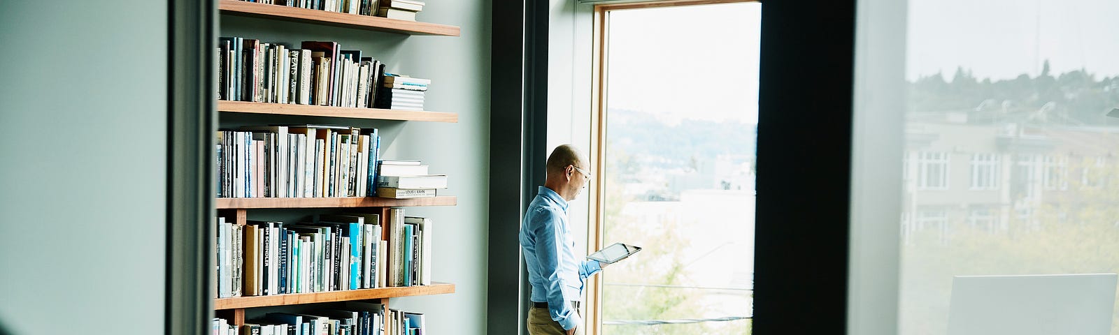 A businessman reading on his digital tablet while standing near a window, next to his filled bookshelves.