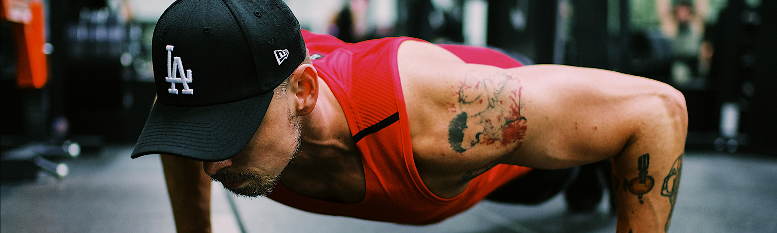 Man in gym wearing a red tank top and black cap, doing a pushup