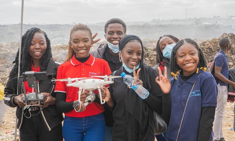 Five girls standing with drones in front of a rocky beach background. Two on the right and a boy behind them are giving peace signs.