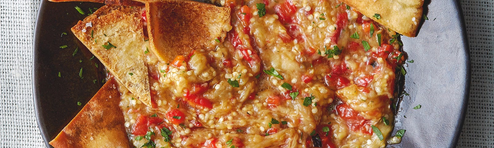 A wide dinner bowl of eggplant dip with toasted pita chips on it.