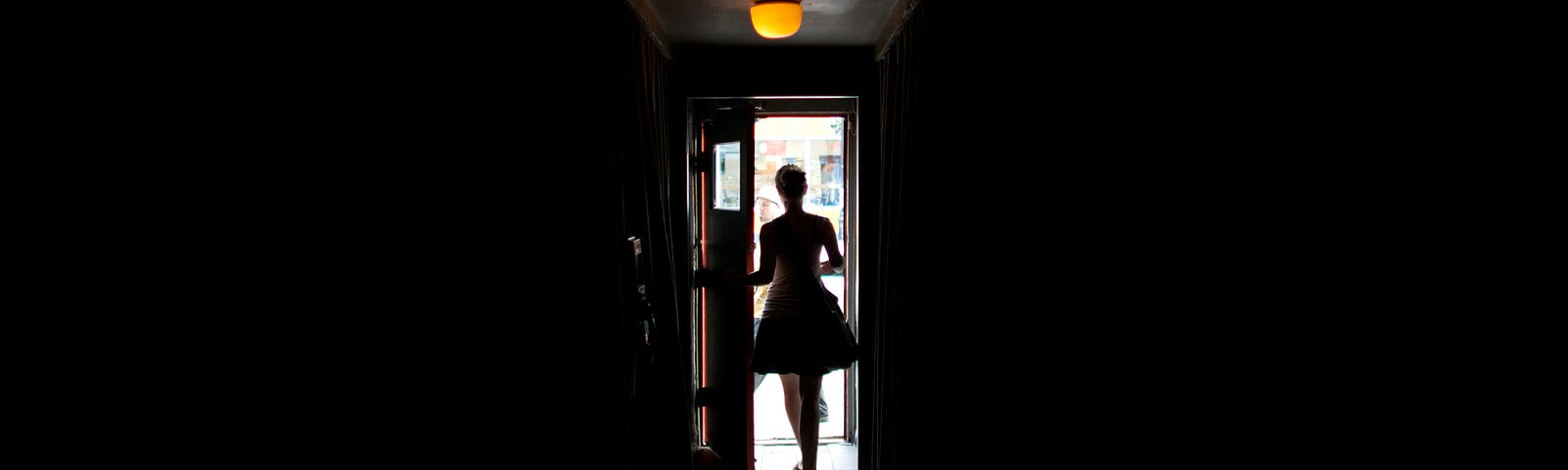 A mostly dark photo of a woman who is opening a door that casts light in the middle of the picture.
