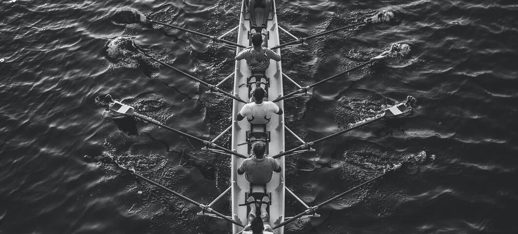 A black-and-white picture of 4 male rowers plus a coxswain in a sleek rowboat