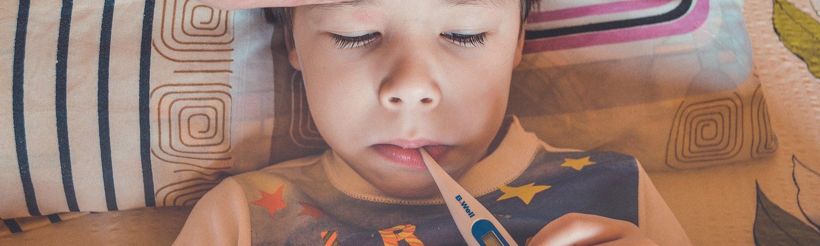 Child in bed with thermometer. Mom’s hand on his forehead