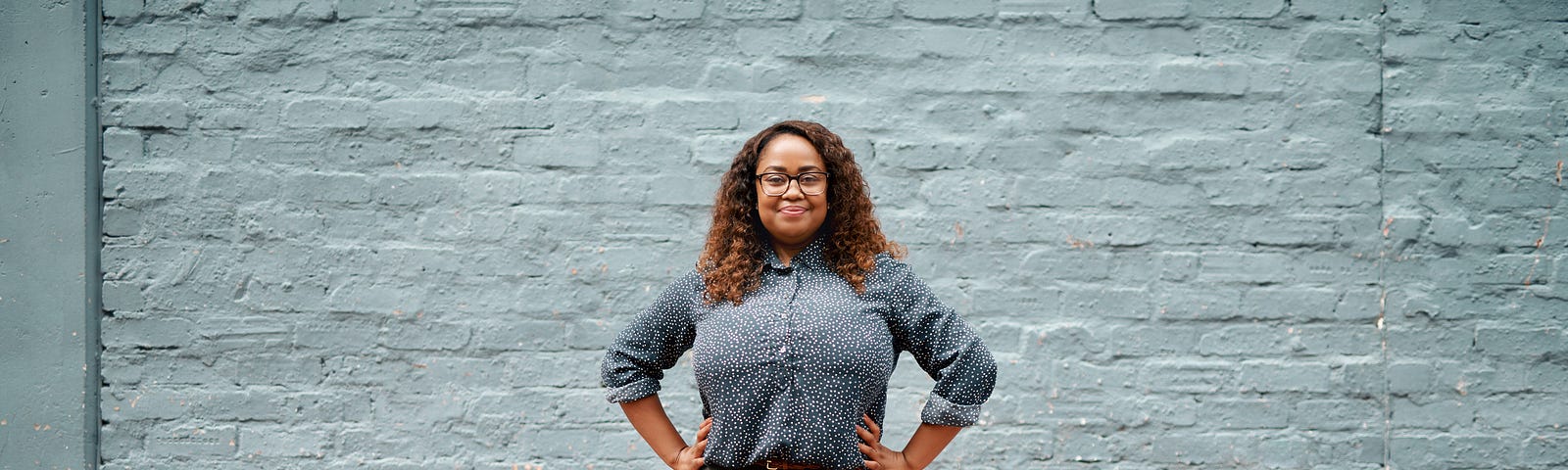 A photo of a confident black woman standing against a blue brick background in a power pose.