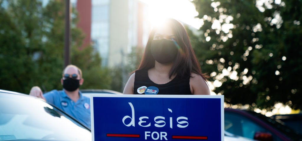 A woman wearing face mask holds a sign that says “desis for Biden Harris.”