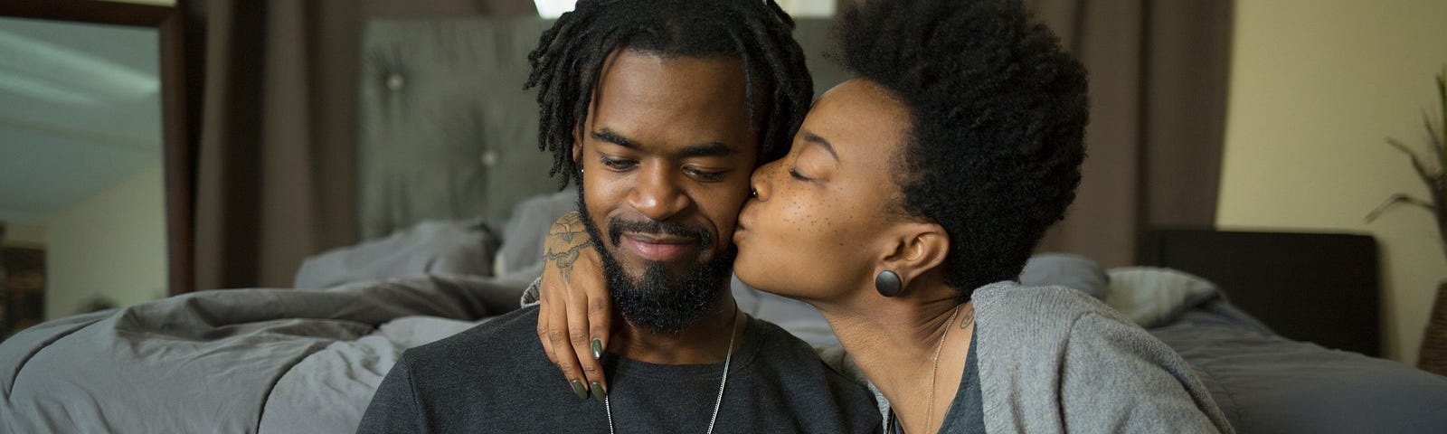 A photo of a black couple. He is playing keyboard and she kisses him on the cheek.