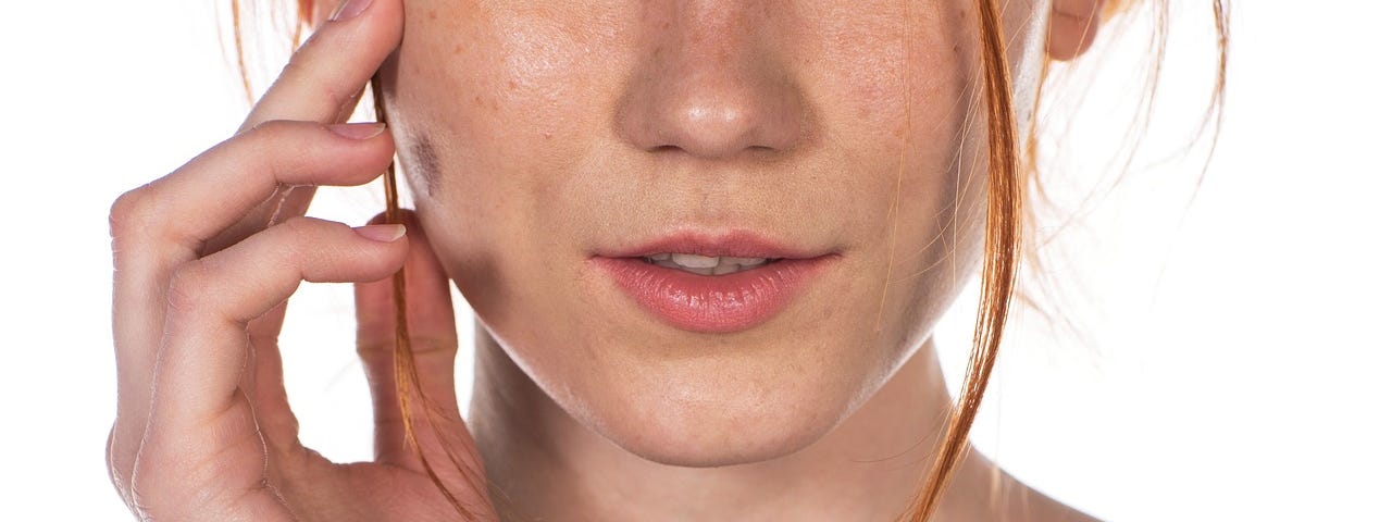 Young woman, model, red hair, ginger tied back, freckles, wears black knit top with boat neck, raises hand to touch her face