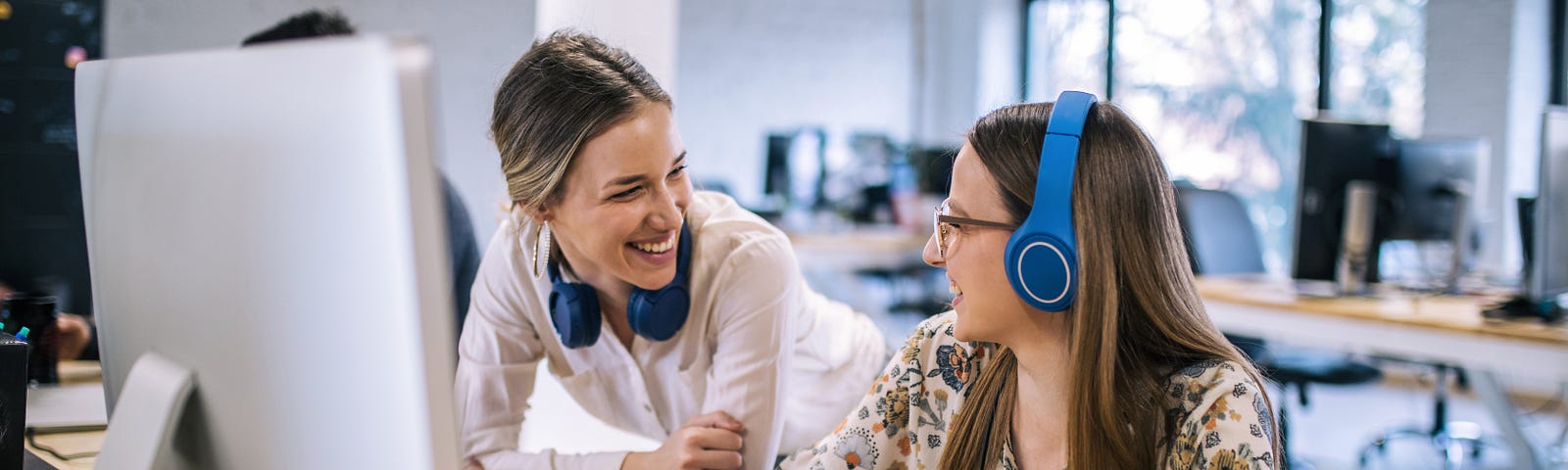 An employee working at her desk with headphones in is interrupted by a manager who comes by for a chat.