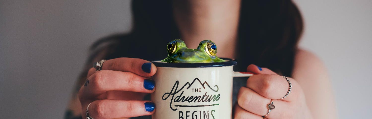 A tattooed woman holds a coffee mug with “The Adventure Begins” written on it. A frog is peeking out of the top of the mug.