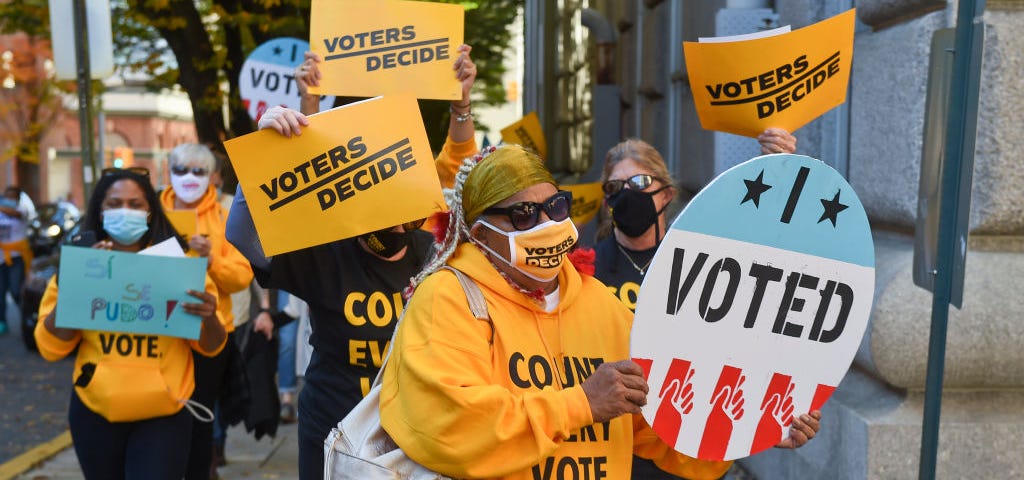 Masked protestors holding signs that say “VOTERS DECIDE” and “I VOTED.”
