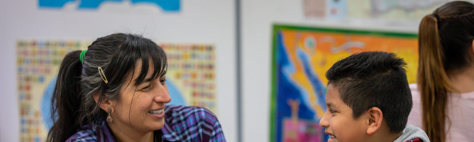 A female teacher talks to a young male student. Both are smiling and in a classroom.