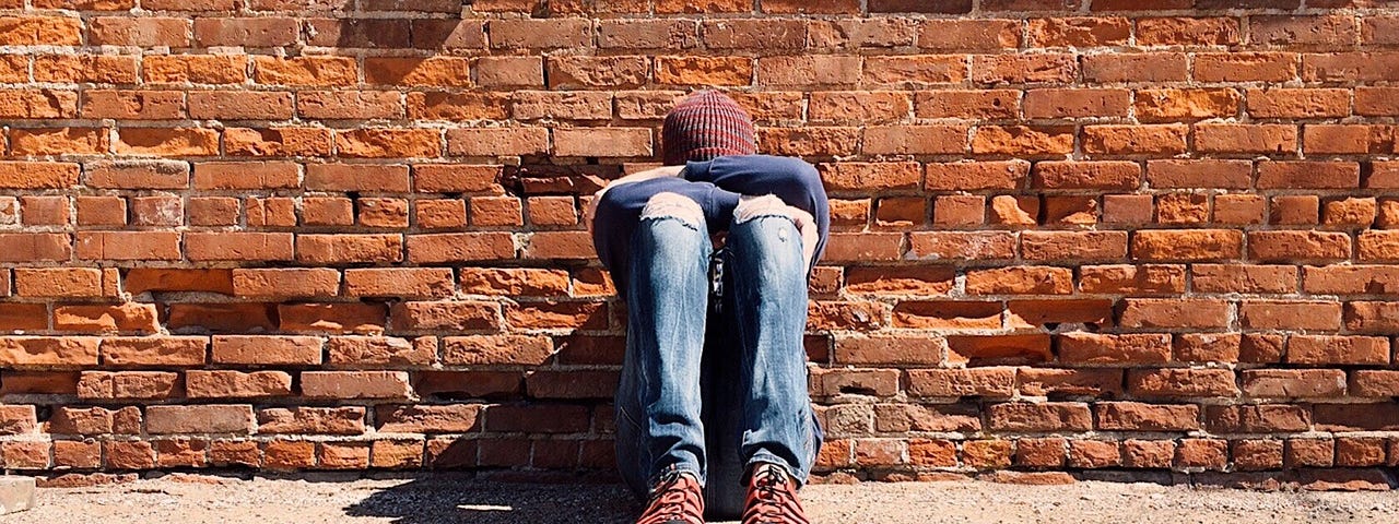 A person in blue jeans. brown boots and beanie sitting on the ground with their back against a brick wall. They have their feet on the ground and their knees up. Their arms are folded across their knees and their head is resting on their arms so their face is hidden.