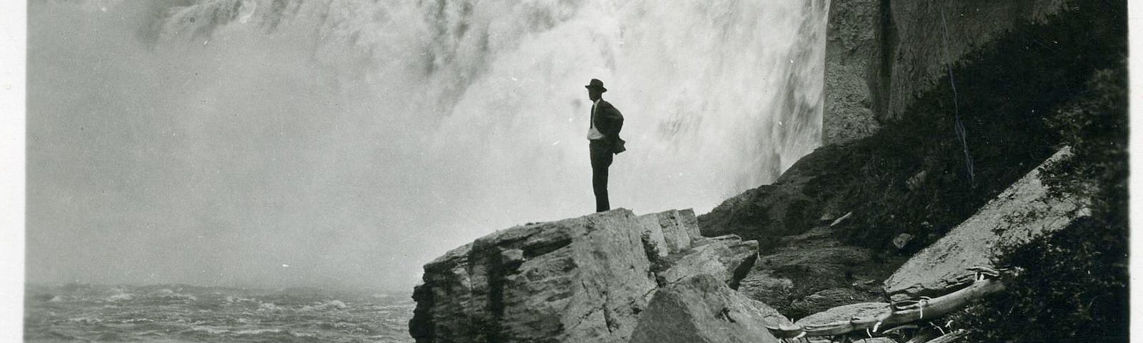 Vintage black-and-white photo of a person standing in front of waterfall.