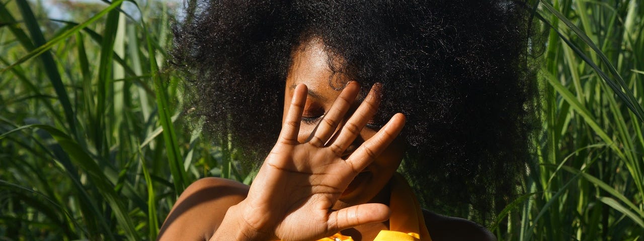Black woman covering face with a hand, standing in a field of tall grass.