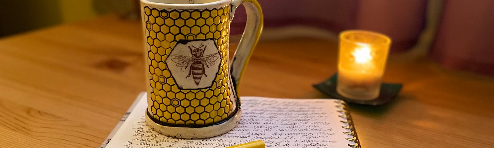 A yellow Lamy pen and a coffee mug with a bee on it, atop a coil notebook filled with cursive writing.