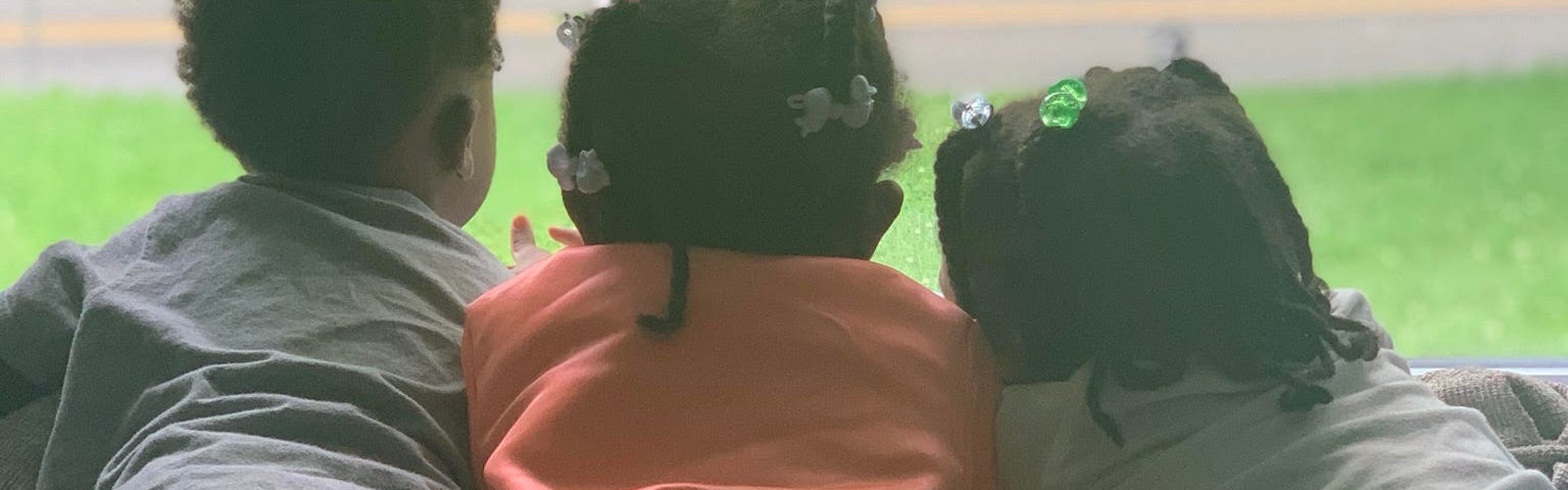 3 young Black children seen from behind, looking out the window at grass and a road.