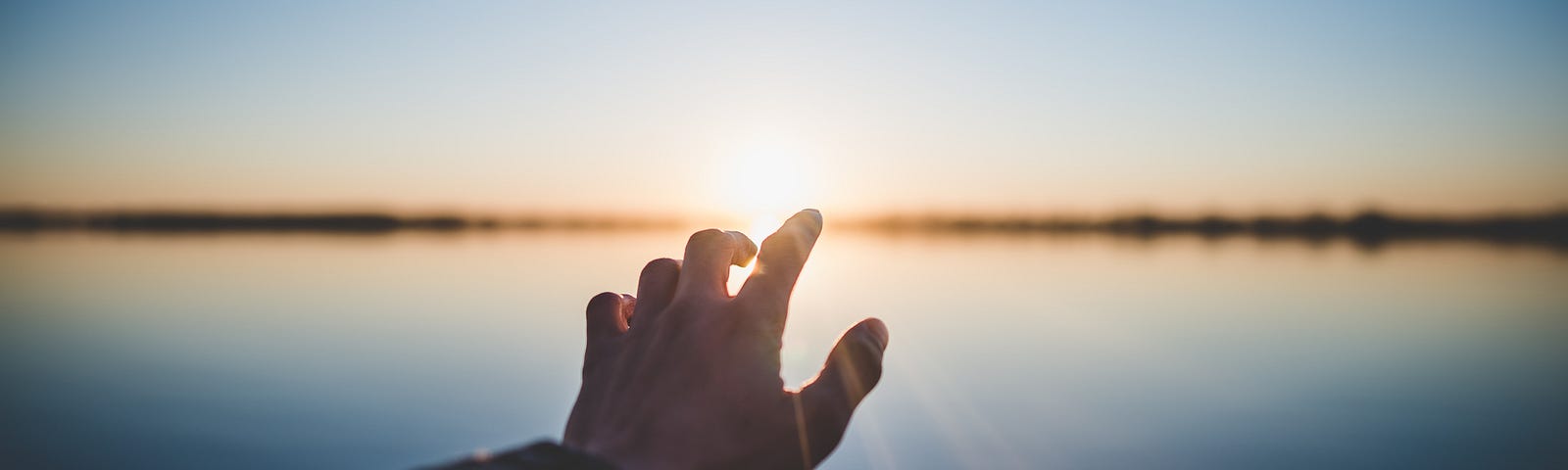 A hand reaching across water for the setting sun.