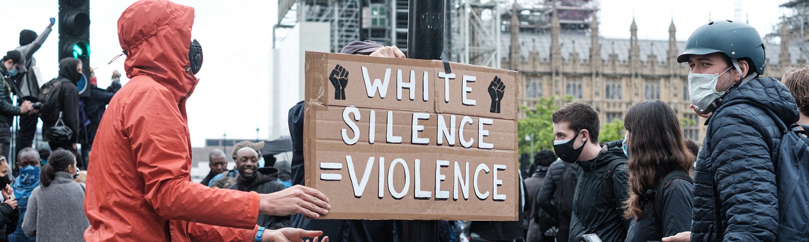 A masked white man holds a cardboard sign while white onlookers watch. The sign says “White Silence Equals Violence.”