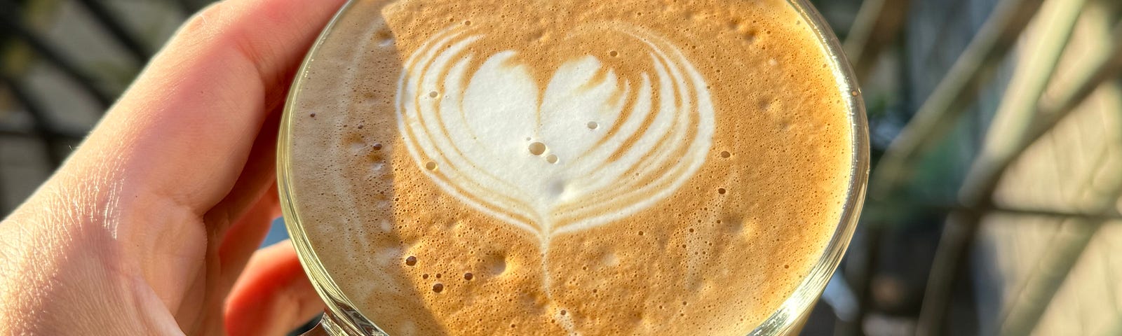 Hand holding a cup of coffee with heart-shape latte art, sun shining, over a garden view from a Juliette balcony.