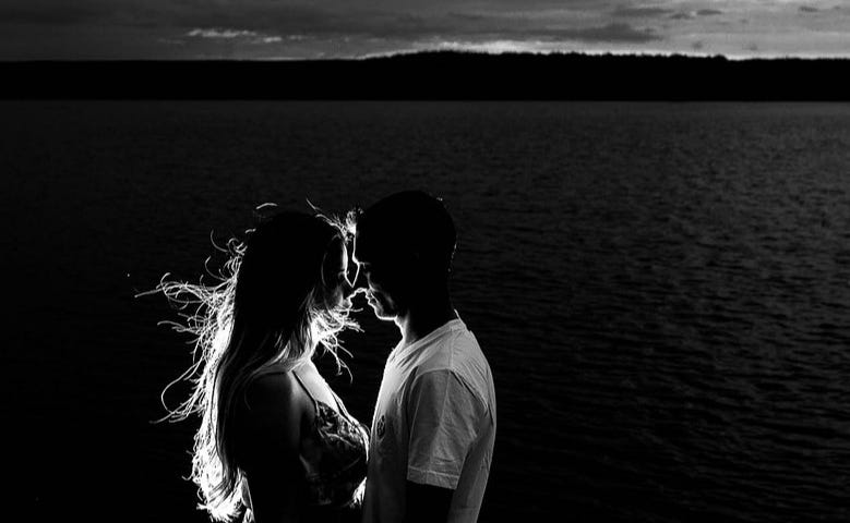 A Monochrome silhouette of a man and woman on a beach in an embrace. Sunlight illuminates the woman’s hair blowing in the wind