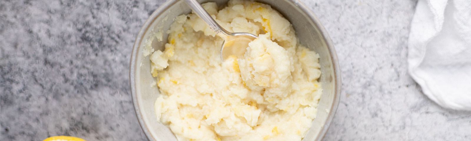 A bowl of mashed potatoes with a spoon on it, next to some lemon slices on a marble countertop.