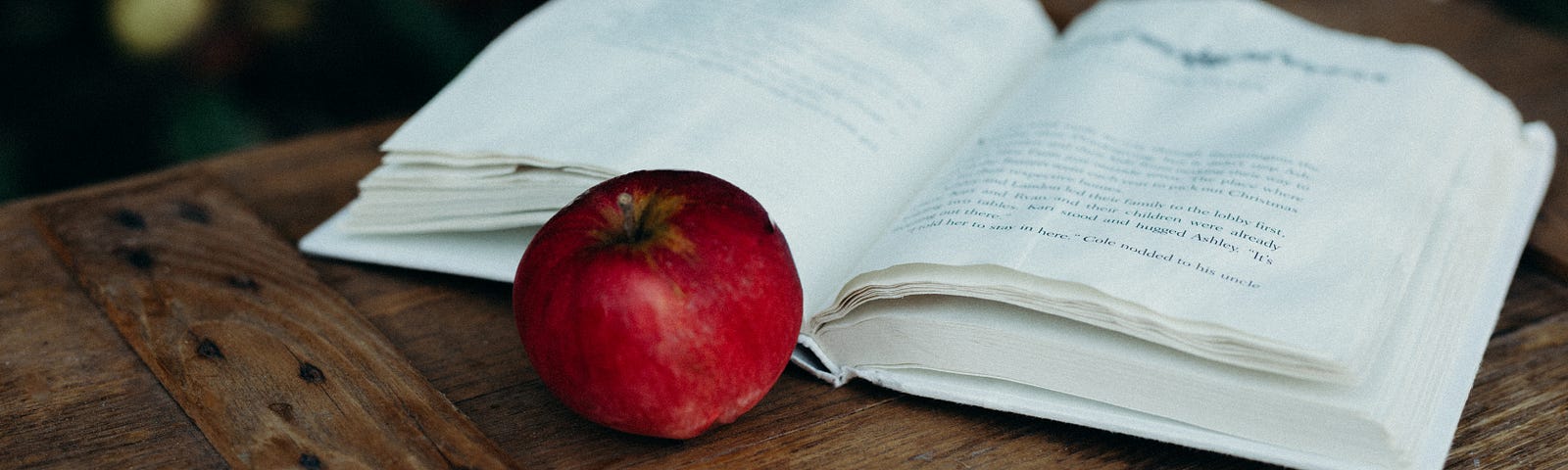 A bright red apple sits in front of the wrinkled pages of an open book. The book and apple sit on top of a wooden board in front of a dark and blurry background.