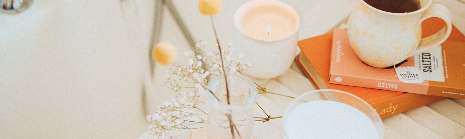 Book, cup of tea, candles, and decorative flower next to a bathtub.