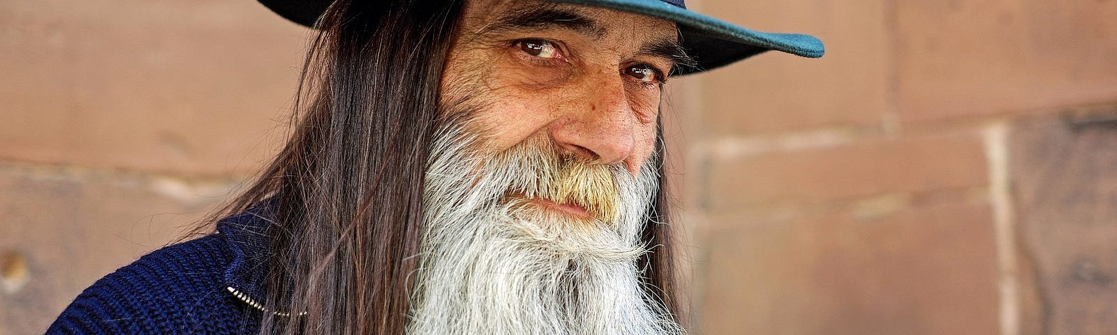 Happy older man with long white beard and long brown hair wearing a teal hat.