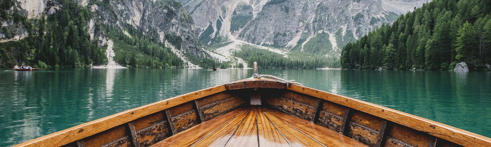 A view of mountains, forest, and a lake, from the position of someone seated in a wooden boat on the lake.