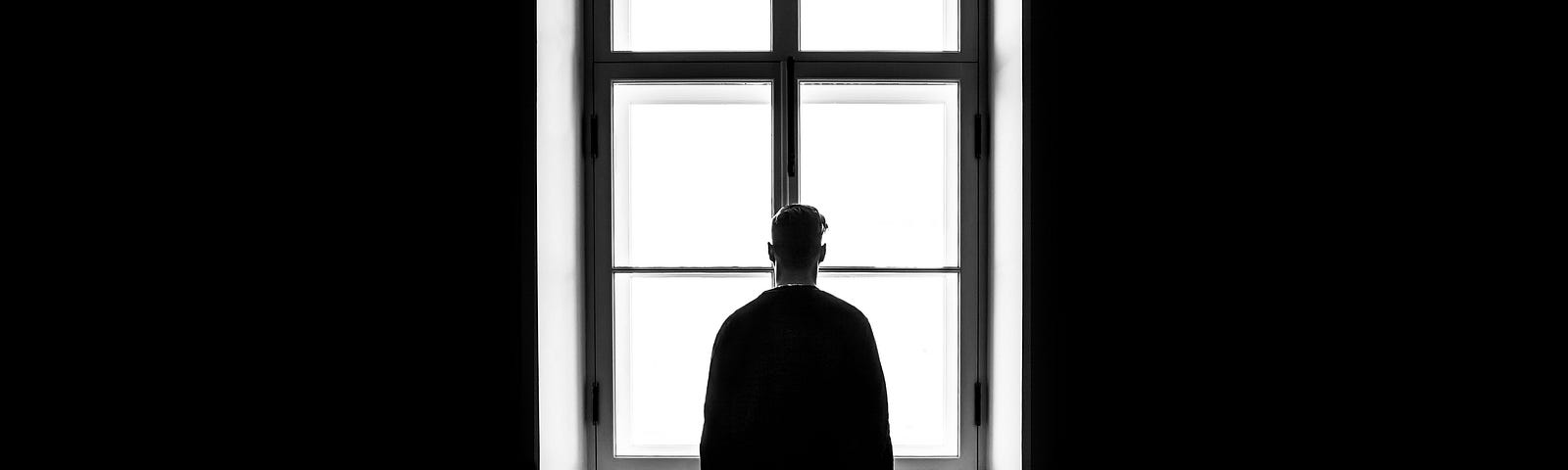 The silhouette of a young man looking outside a window alone.