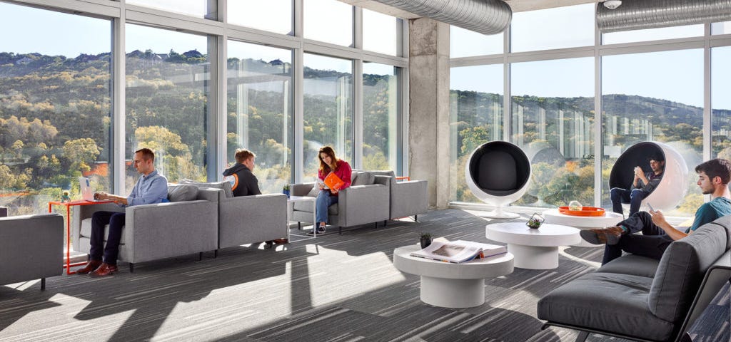 An open office lounge area with multiple people sitting down and using laptops or mobile phones.