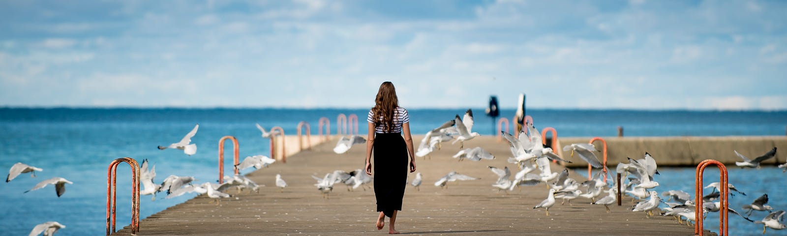 A woman walking on a boardwalk with a beautiful blue sky and ocean in front of her.