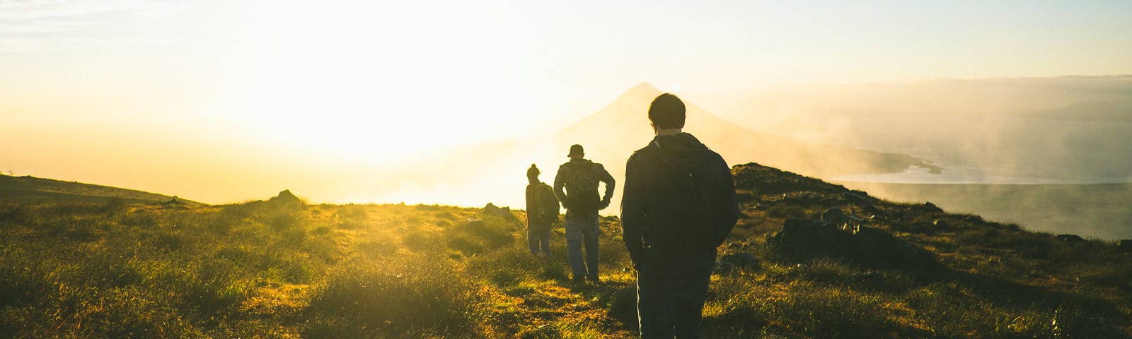 An image of three hikers walking up a mountain towards the sun.