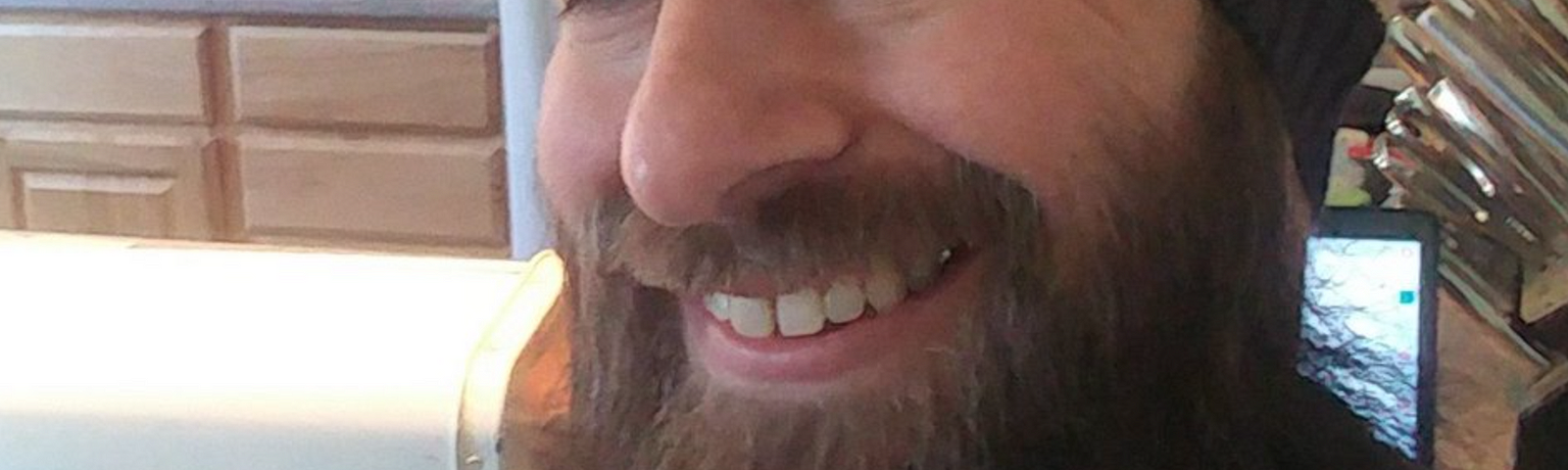 Photograph of the author, Hogan Torah, grinning, wearing beige and brown striped beanie hat. Full beard. Appears to be in kitchen.