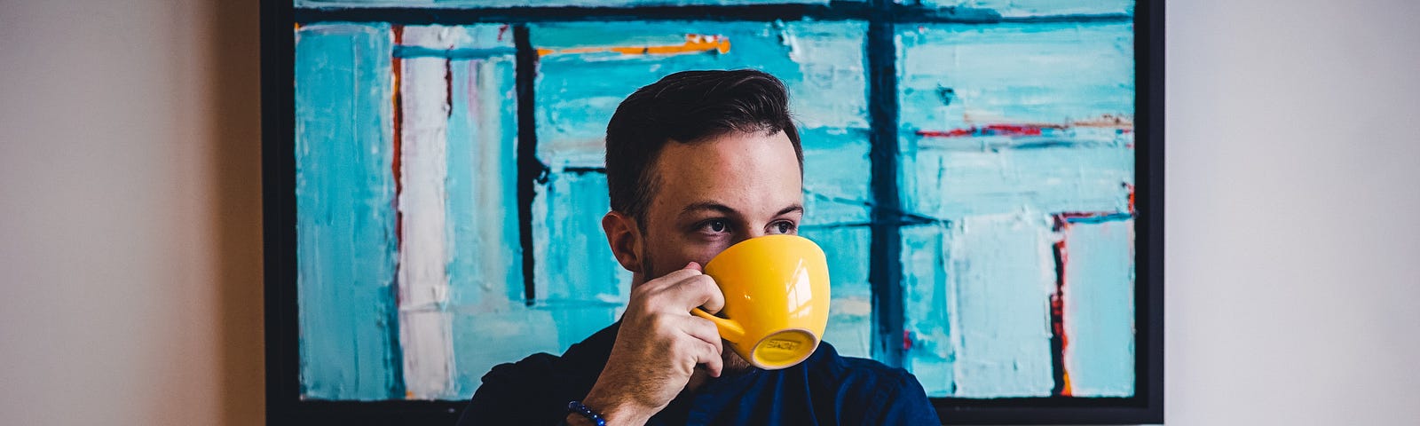 Man sips from a yellow mug while using a laptop. Angular oil painting on the wall behind him.