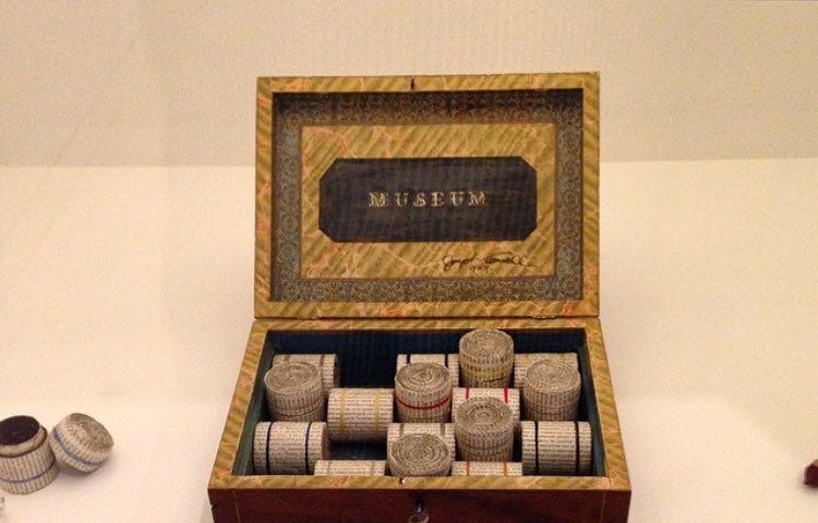 A small wooden box with its lid open. Inside the lid is a label that simply says “MUSEUM”. Inside the box are a number of small cylinders which look to be made out of either thing wood or cardboard. On the shelf outside the box, several similar cylinders are open with their contents viewable, featuring small objects like paper clips, string, etc.