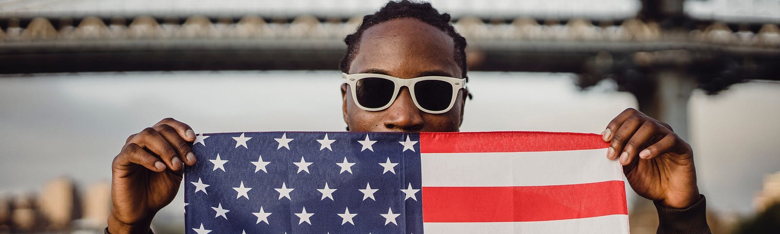 Black man wearing sunglasses, holding American flag up with both hands. Flag covers his mouth.